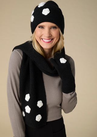 A smiling blonde woman wearing a black knit beanie, matching scarf, and knit mittens with white polka dots, posed against a solid beige studio background.