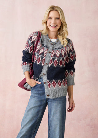 A smiling blonde woman models a cozy knit cardigan with a navy, gray, and burgundy Fair Isle diamond pattern, paired with blue jeans and a burgundy shoulder bag against a soft pink background.
