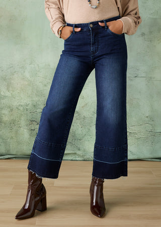 A person wearing dark blue wide-leg cropped jeans with contrast stitching and raw hems, styled with a beige long-sleeve top and dark brown pointed-toe heeled boots against a textured green wall and light wood floor.
