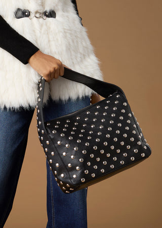 A person wearing a white faux-fur vest, black top, and blue jeans holds a black studded shoulder bag against a solid tan background.
