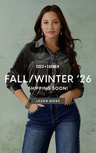 A woman stands against a textured light green wall wearing a black and gray striped button-up shirt and blue jeans with silver stud detailing on the pockets. Large white text overlays read 'COCO + CARMEN', 'FALL/WINTER '26 SHIPPING SOON!', and 'LEARN MORE'.