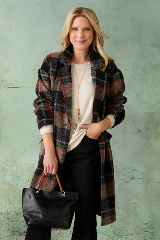 A woman models a long, open-front plaid coat in earthy tones of brown, black, and red, worn over a cream top and black pants, holding a black tote bag against a textured sage green backdrop.