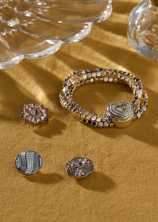 A jewelry set displayed on a textured mustard-yellow fabric background, featuring a multi-strand beaded bracelet with a heart-shaped clasp and three matching stud earrings with intricate crystal and metallic detailing.