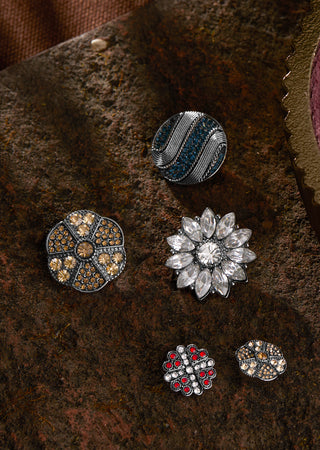 Four ornate jewelry pieces arranged on a textured dark brown surface: a round silver-toned piece with blue and silver wavy detailing, a round silver-toned piece with gold and brown stones, a silver-toned flower-shaped brooch with clear stones, and two small round pieces with red and gold stones.