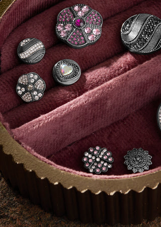 A close-up of several ornate jewelry charms or button-like accessories arranged inside a round velvet jewelry box. The pieces feature silver-toned metalwork with pink and clear rhinestones, and one central piece has a heart-shaped clear stone.