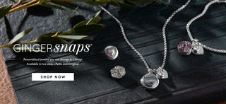 Silver snap chain necklaces with heart and oval charms, displayed on a dark textured surface with green leaves. The brand name 'GINGER snaps' is visible on the left with a 'SHOP NOW' button.