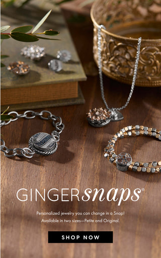 A styled product display of personalized jewelry featuring silver and gold-toned bracelets, a necklace, and earrings on a rustic wooden table with decorative props. The text 'GINGERSnaps' is prominently displayed with a 'SHOP NOW' button below.