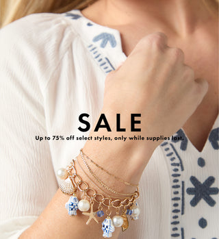 A close-up of a woman wearing a delicate gold charm bracelet with sea-themed charms including a seashell, starfish, and blue-and-white fish figures, paired with a white embroidered blouse. Large text overlay reads 'SALE' with smaller text below.