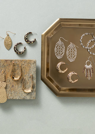 A styled flat-lay of multiple pairs of earrings displayed on a metallic gold tray and a textured stone block against a pale blue-gray background.