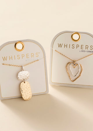 Two gold-tone Whispers necklaces on display cards against a warm cream background. One necklace has a textured white bead and a hammered gold tag pendant, and the other has a double-heart outline pendant.