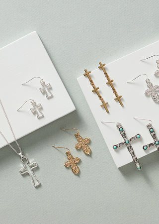 A styled flat-lay of multiple cross-shaped jewelry pieces, including earrings and a necklace, displayed on white cards against a pale blue background.