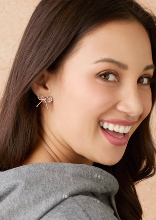 A close-up of a woman with long dark brown hair smiling, wearing a small bow-shaped stud earring with sparkling clear stones on a warm beige background.