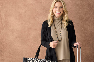 A smiling blonde woman stands against a textured brown backdrop, wearing a black cardigan over a taupe turtleneck and holding a black-and-white patterned tote bag and a rolling suitcase.