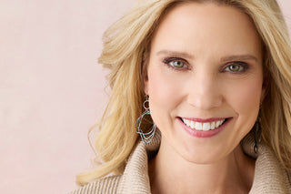 A close-up portrait of a smiling blonde woman wearing large silver-toned geometric dangle earrings and a beige knit top against a soft pink background.