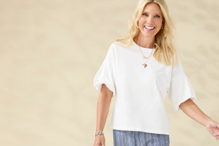 A smiling blonde woman wearing a loose white short-sleeve top and blue striped pants, standing against a soft beige background.