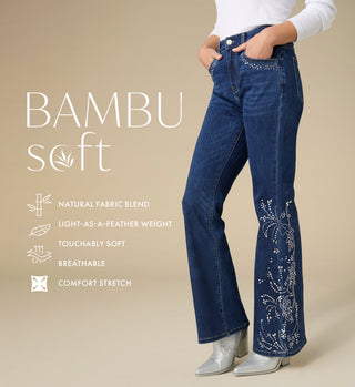 A model wears dark blue bamboo-soft flare jeans with white floral embroidery and rhinestone embellishments on the lower legs, paired with a white long-sleeve top and silver pointed boots. The image features a beige background with text highlighting the product's bamboo fabric blend, light-as-feather weight, touchably soft texture, breathability, and comfort stretch.