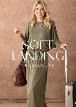 A woman modeling an olive green long-sleeve maxi dress with a gathered waist, standing against a textured brown backdrop and holding a suitcase and a brown bag. Large text overlay reads 'SOFT LANDING TRAVELWEAR'.