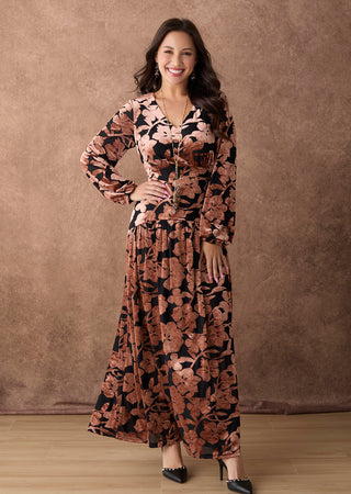A woman modeling a long-sleeve maxi dress with a brown and black floral pattern, standing against a textured brown backdrop.