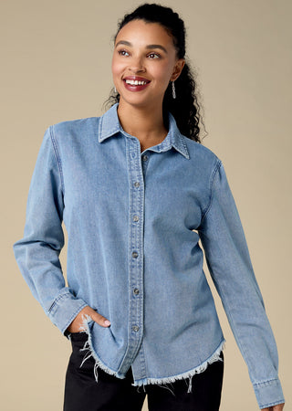 A woman wearing a light blue denim button-up shirt with a collar and raw frayed hem, paired with black pants, against a solid beige studio background.