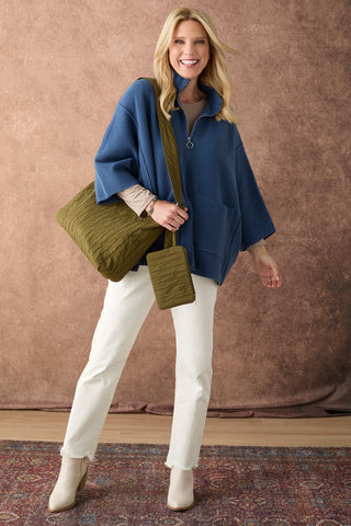A woman models a blue quarter-zip pullover, white pants, and a large olive green quilted crossbody bag against a textured brown backdrop.