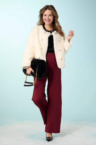 A woman models a stylish outfit consisting of a cream faux-fur cropped jacket, a black lace-trimmed top, burgundy wide-leg pants, and a black faux-fur clutch bag with a gold chain strap, set against a light blue studio background.