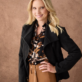 A woman modeling a black suede jacket over a brown and black patterned blouse, paired with brown pants, set against a textured brown studio backdrop.