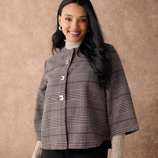 A woman wearing a brown and gray plaid cropped jacket with wide sleeves over a cream mock-neck top, standing against a textured brown backdrop.
