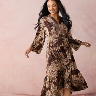 A woman modeling a brown and beige floral print midi dress with long bell sleeves and a tiered ruffled hem, set against a solid dusty pink background.