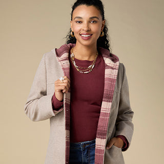 A woman modeling a light beige hooded cardigan with a dusty rose and beige plaid lining, worn over a burgundy top and blue jeans against a solid beige background.
