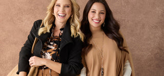 Two women smiling and posing together against a textured brown studio backdrop. The woman on the left wears a black jacket over a patterned top, and the woman on the right wears a tan long-sleeve top with a long gold necklace.