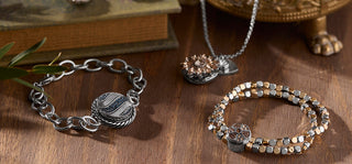 A styled flat-lay of three jewelry pieces on a wooden surface: a silver chain bracelet with a round textured charm, a silver necklace with a flower-shaped pendant, and a beaded stretch bracelet in silver, gold, and rose gold tones with a central charm.