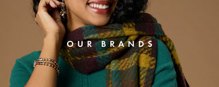 A close-up lifestyle shot of a smiling woman wearing a green sweater and a dark green, brown, and mustard plaid scarf. She is also wearing gold bracelets and earrings.