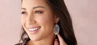 A close-up of a smiling woman wearing a dangling silver filigree earring against a soft pink background.
