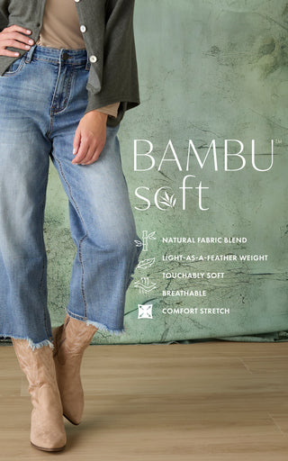 A person wearing light-wash blue cropped denim jeans with a raw hem, styled with beige boots and a dark cardigan, standing in front of a textured green wall with text reading 'BAMBU soft'.