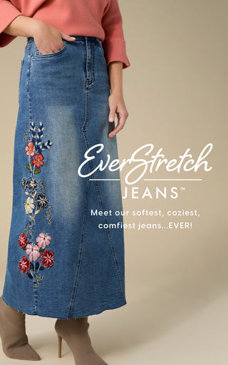 A model wears a blue denim midi skirt with colorful floral embroidery down the side, paired with a coral top and beige ankle boots. The image features a beige background with a text overlay that reads 'EverStretch JEANS' and 'Meet our softest, coziest, comfiest jeans.