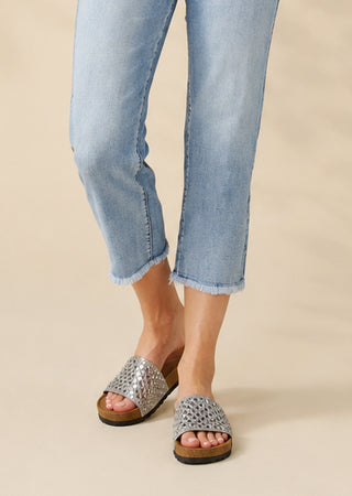 A person wearing light blue cropped denim jeans and silver woven slide sandals with brown soles, shown from the knees down against a plain beige studio background.