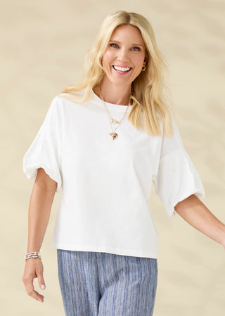 A smiling blonde woman wearing a loose white short-sleeve top with voluminous elbow-length sleeves and blue striped pants, standing against a soft beige studio background.