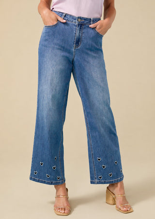 A person wearing blue wide-leg cropped jeans with small embroidered heart details near the hem, paired with a light pink top and beige heeled sandals, against a plain beige studio background.