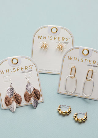 Gold and silver earrings on 'Whispers' branded cards against a light blue background